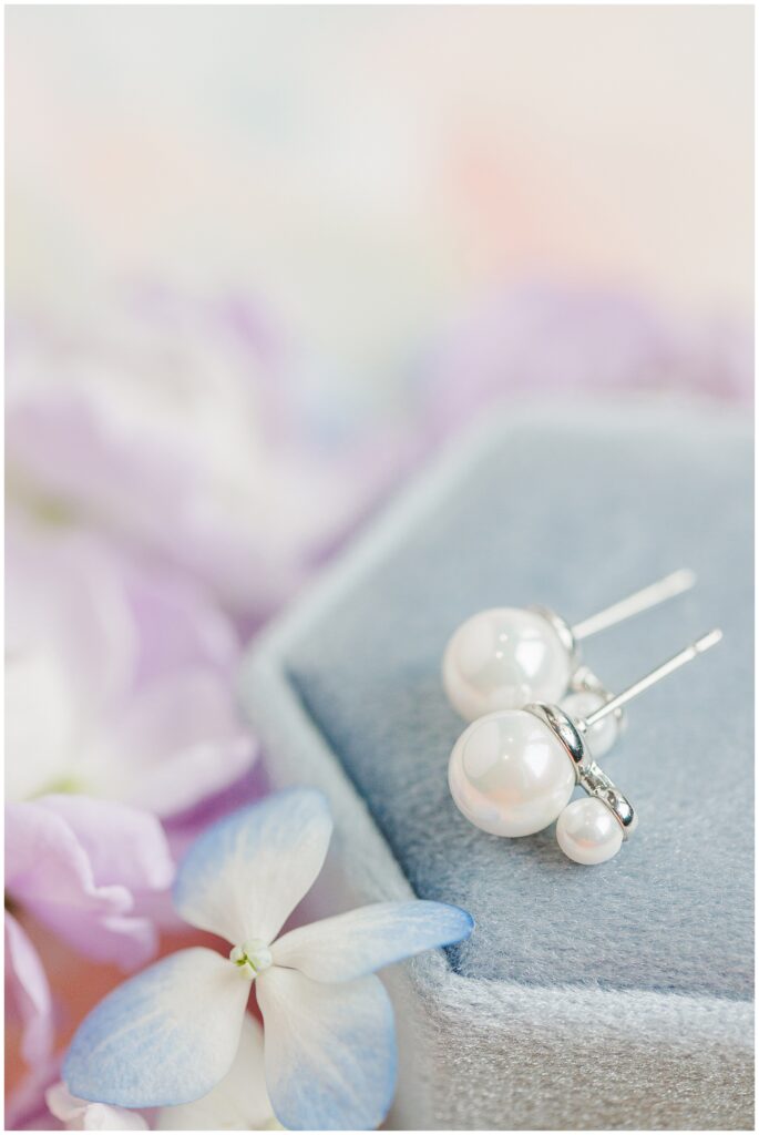 A pair of pearl earrings next to a blue hydrangea flower, resting on a soft blue velvet surface.