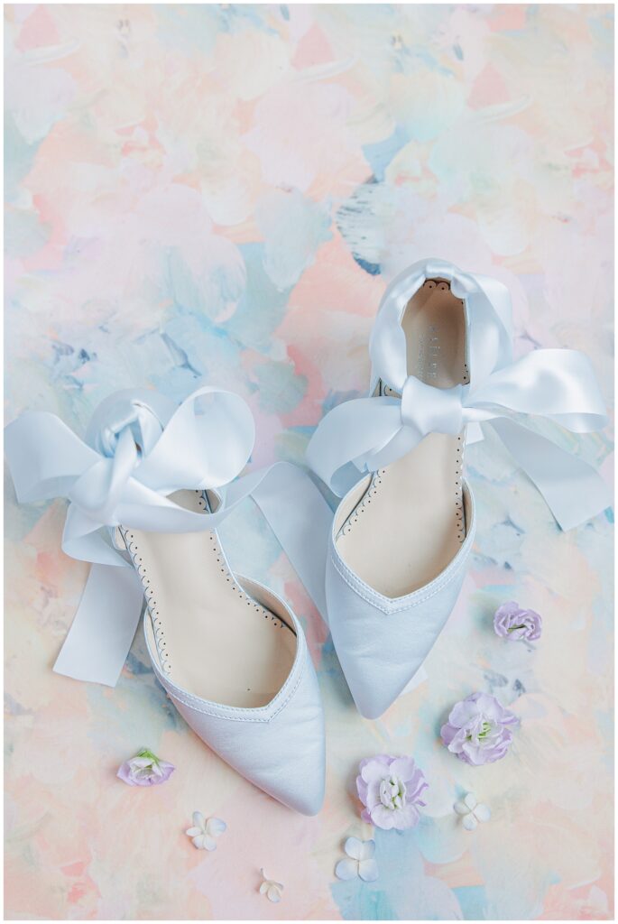 A pair of blue bridal shoes with pointed toes and satin ribbon ties, placed on a pastel floral background with scattered purple and white flowers.