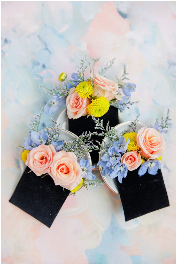 Three floral corsages featuring peach roses, yellow blooms, and blue flowers, displayed against a similar pastel background.