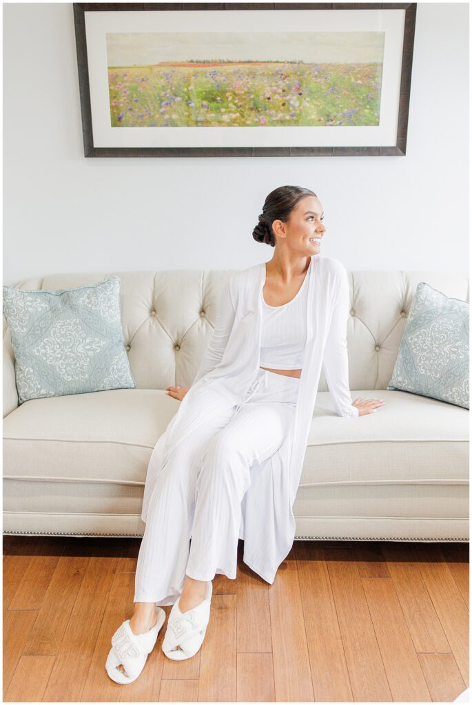 A bride wearing a white loungewear set, sitting on a cream-colored sofa and smiling, with her hair styled in an updo. A floral painting is hung on the wall behind her.