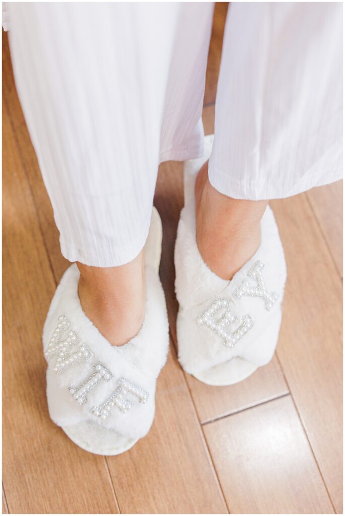 A close-up of white fuzzy slippers with the word “WIFEY” spelled out in pearl embellishments, worn by the bride.