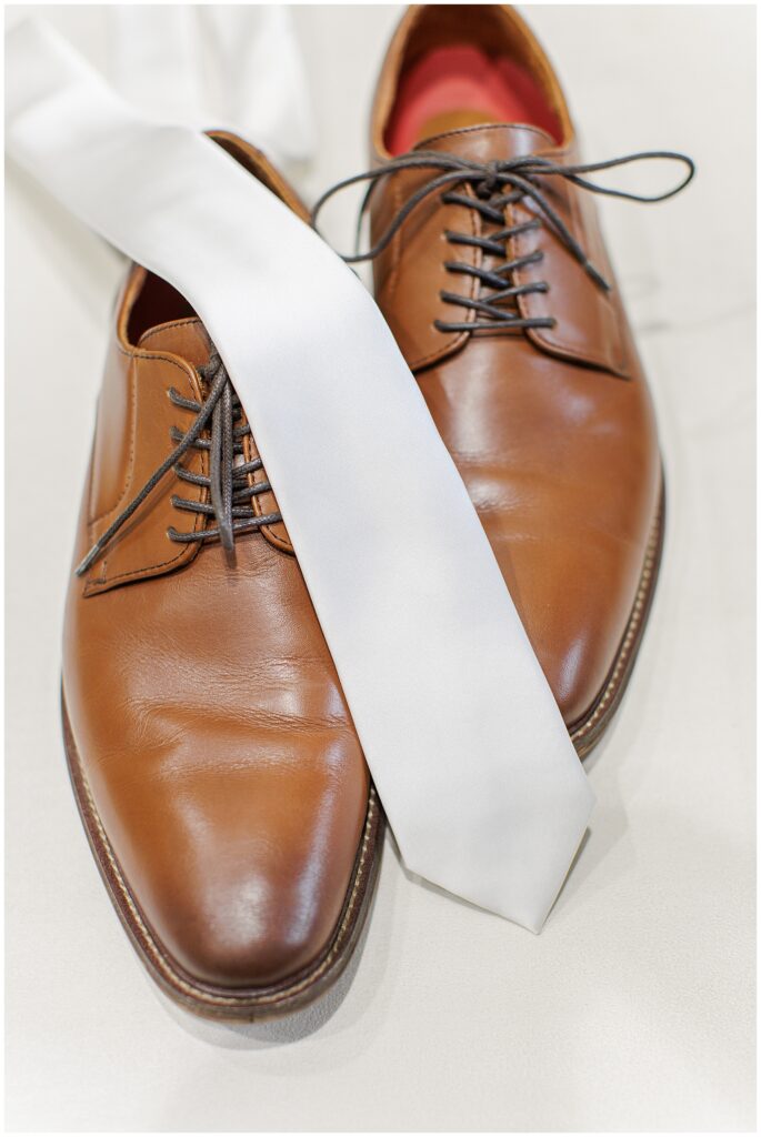 A pair of brown leather dress shoes with a white necktie draped across them.