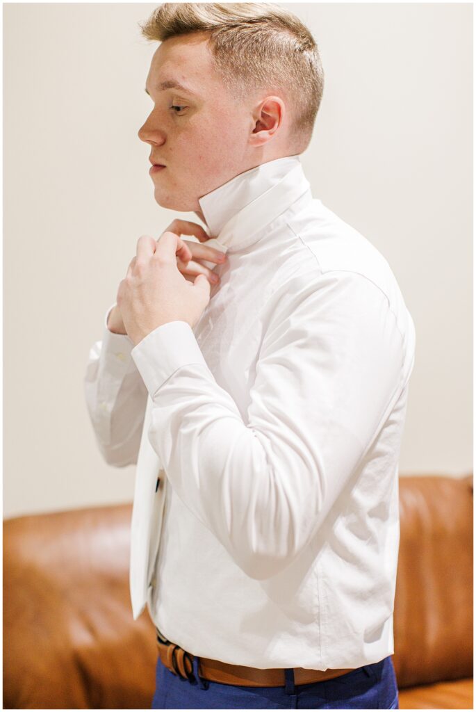 A groom adjusting his white dress shirt collar, preparing to tie his necktie.