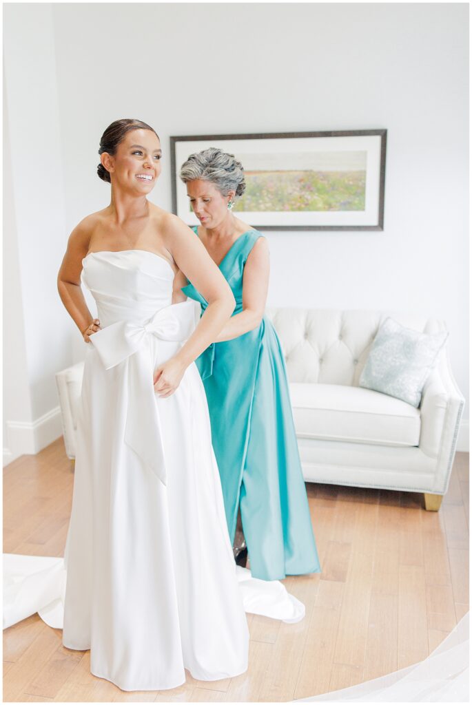 The bride smiling as her mother, dressed in a teal gown, helps adjust her white wedding dress with a large bow on the waist.