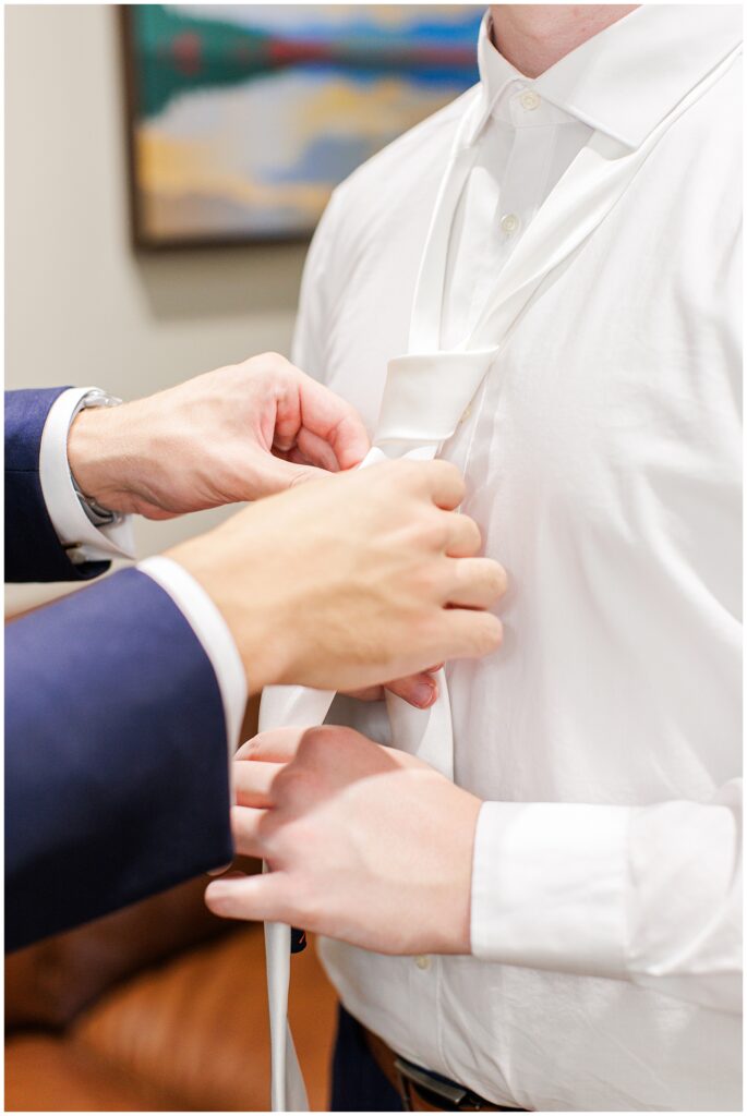 A groomsman tying the groom’s white necktie while the groom stands wearing a white shirt and navy blue pants.