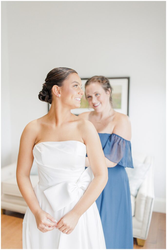 The bride smiling as a bridesmaid in a navy blue dress helps her get ready, with both standing in a brightly lit room.