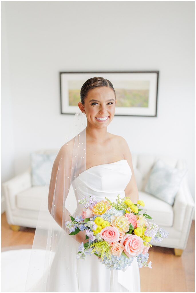 Close-up of the smiling bride in a white wedding dress with a pearl-embellished veil, holding a pastel-colored bouquet of roses and hydrangeas. Taken at Pembroke Pines Country Club, NH.