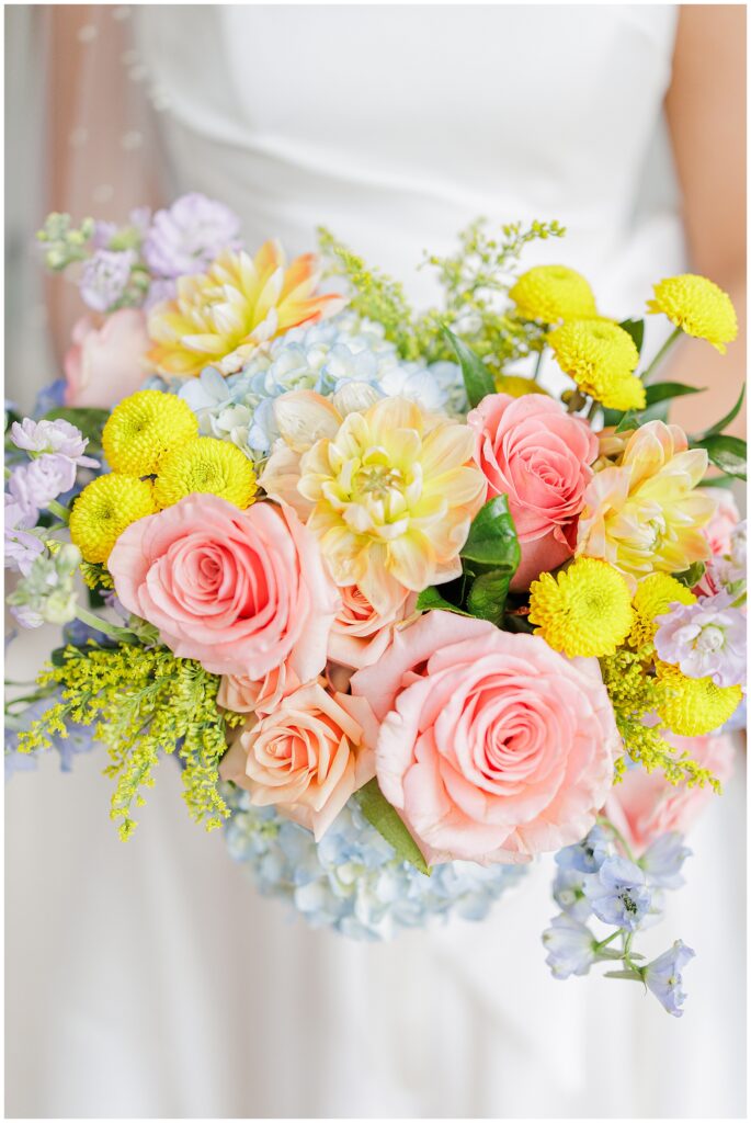 Detailed shot of the bride’s bouquet featuring pink roses, yellow dahlias, blue hydrangeas, and yellow accents. Captured at Pembroke Pines Country Club in Pembroke, NH.