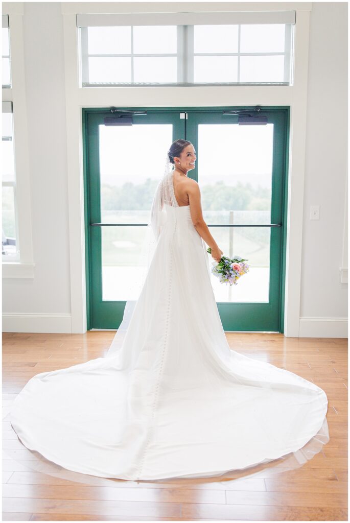 Bride standing in front of large windows at Pembroke Pines Country Club, her white gown trailing on the floor. She holds her bouquet while looking back and smiling.