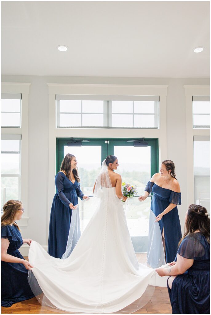 Bride standing near large windows at Pembroke Pines Country Club, with four bridesmaids in navy dresses helping arrange her dress train and veil.