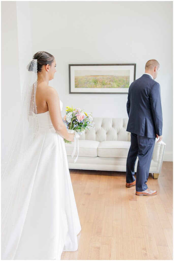 The bride standing behind her father for a first look moment in a bright room at Pembroke Pines Country Club. The father wears a navy suit and the bride holds her bouquet.