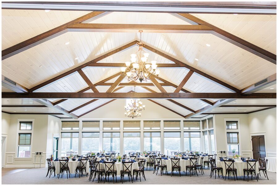 Spacious event hall at Pembroke Pines Country Club, set up for a wedding reception. The room has exposed wood beams, chandeliers, and round tables with navy napkins.