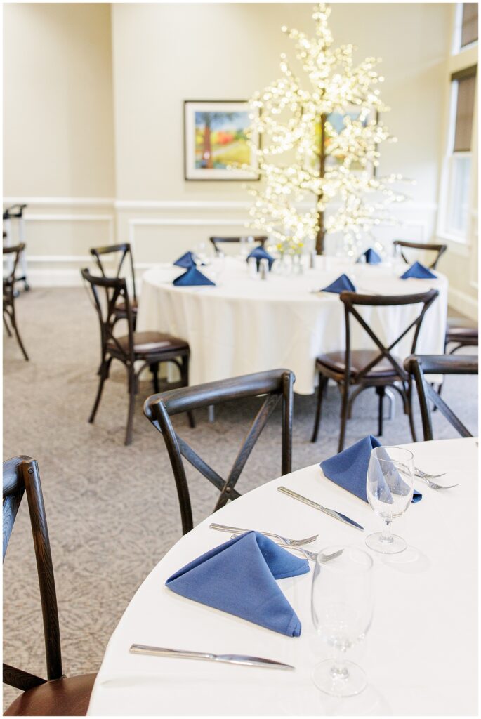 Reception table at Pembroke Pines Country Club, set with navy napkins and glassware, featuring a lighted tree centerpiece in the background.