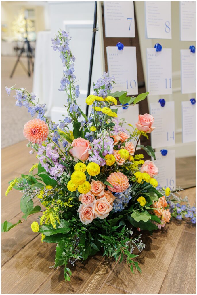 Close-up of the floral arrangement at the base of the seating chart at Pembroke Pines Country Club, featuring peach roses, yellow chrysanthemums, and purple delphiniums.