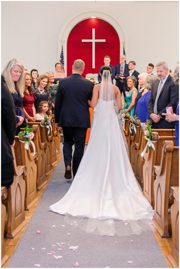 Bride walking down the aisle at a church in Pembroke, NH, with her father. The pews are decorated with flowers, and the altar features a large cross.