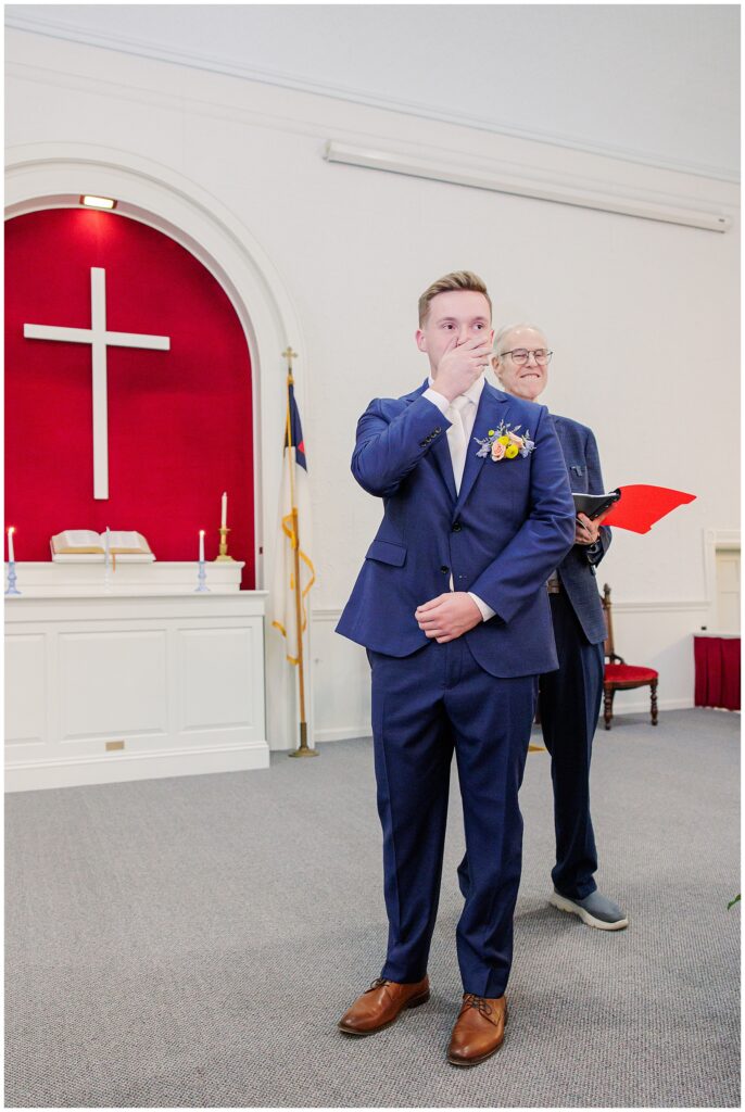 Groom in a blue suit, overcome with emotion, covering his mouth as he watches the bride walk down the aisle at a church in Pembroke, NH.