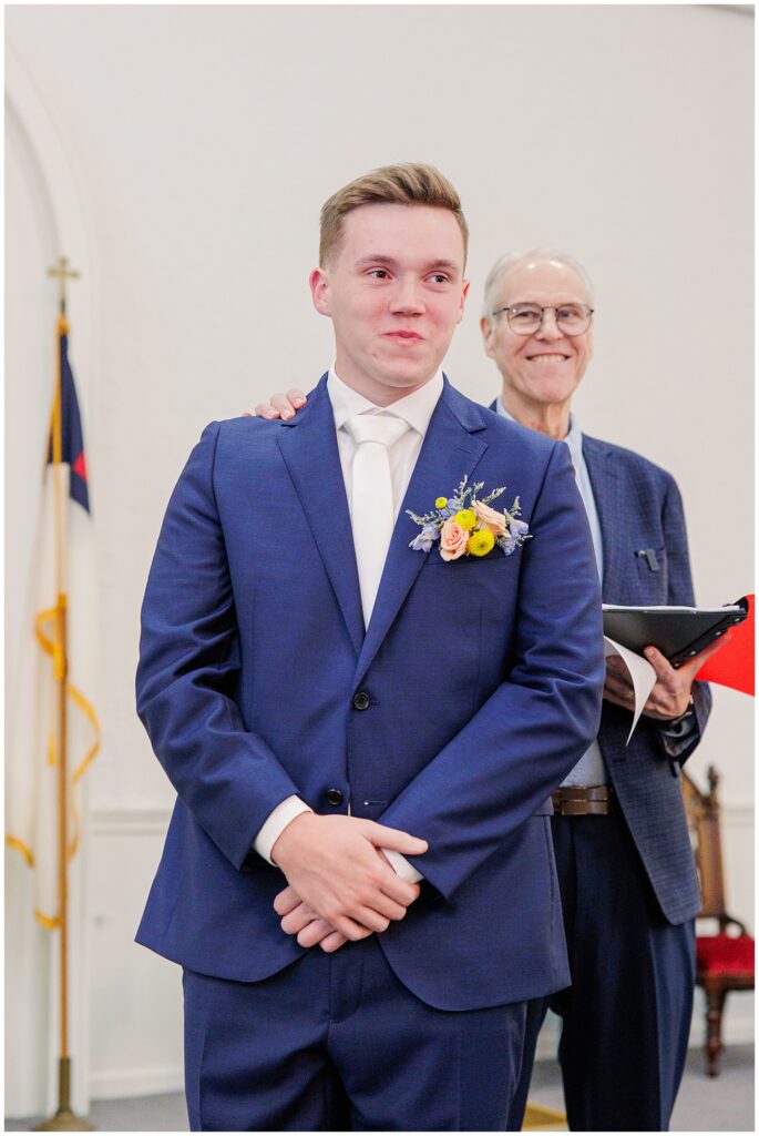 The groom, holding back tears, stands at the altar with the officiant smiling behind him at a church in Pembroke, NH.