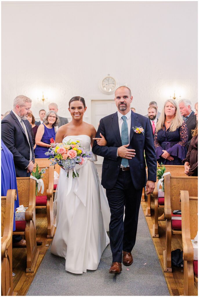 Bride and her father smiling as they walk down the aisle during the ceremony at a church in Pembroke, NH. The bride carries a colorful bouquet of roses and hydrangeas.