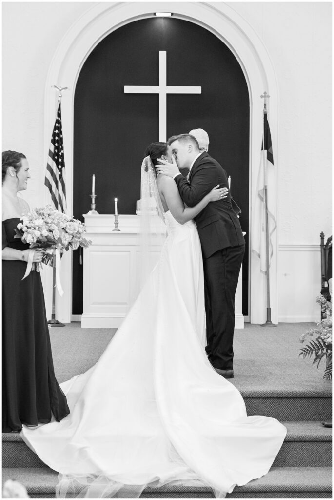 Black-and-white photo of the bride and groom sharing their first kiss at the altar beneath a large cross during their church ceremony in Pembroke, NH.