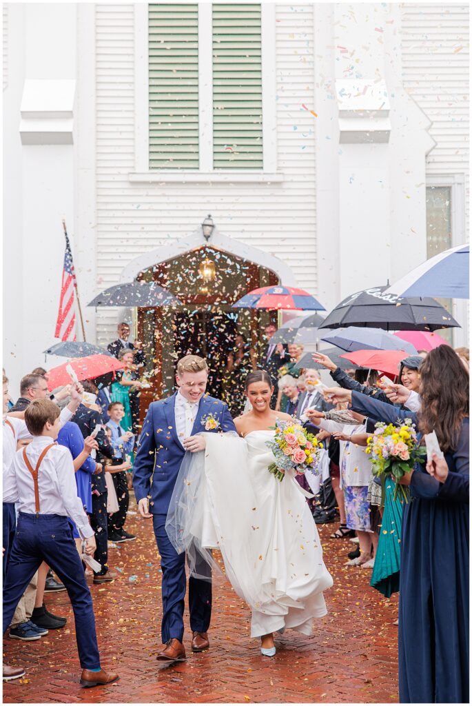 Newlyweds exiting the church in Pembroke, NH, as guests throw confetti. The couple walks under umbrellas on a rainy day, smiling and holding hands.