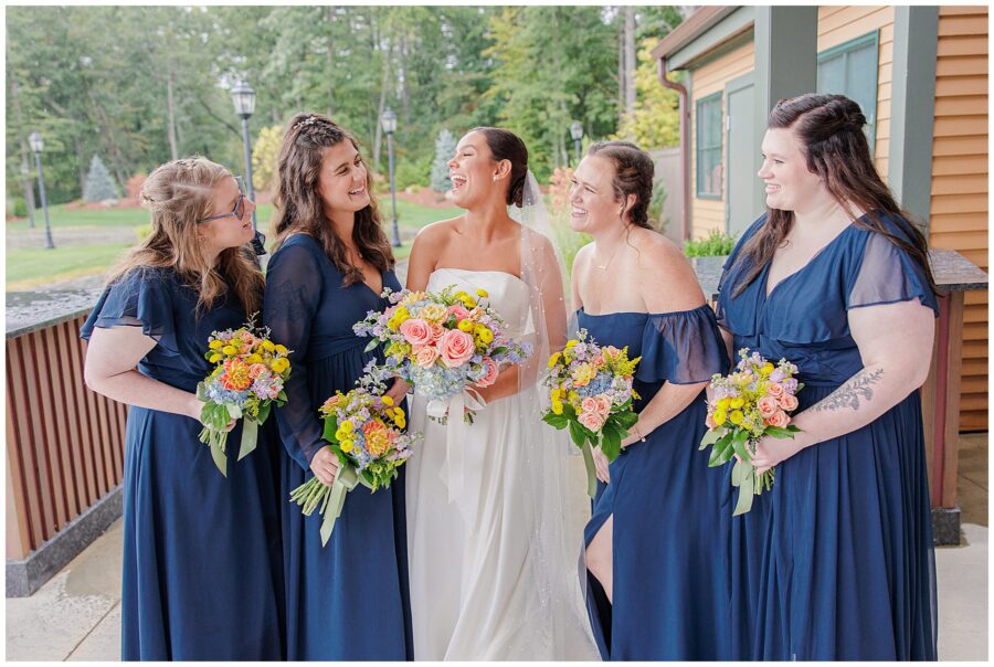 Bride laughing with her bridesmaids at Pembroke Pines Country Club in Pembroke, NH. The bridesmaids wear navy blue dresses and hold colorful bouquets.