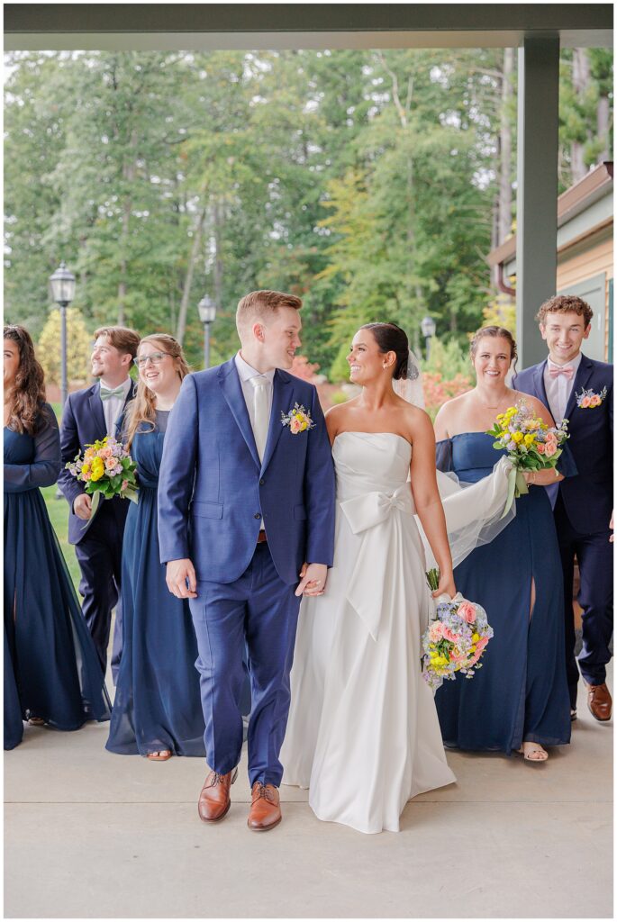 Bride and groom walking with their wedding party under a covered patio at Pembroke Pines Country Club in Pembroke, NH. The bride wears a strapless white gown, and the groom is in a blue suit.