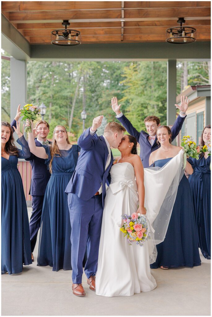 The bride and groom kissing while their bridal party cheers and celebrates under the covered patio at Pembroke Pines Country Club in Pembroke, NH.