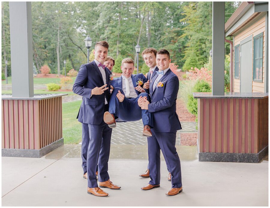 The groomsmen lifting the groom and posing playfully under the covered patio at Pembroke Pines Country Club in Pembroke, NH.