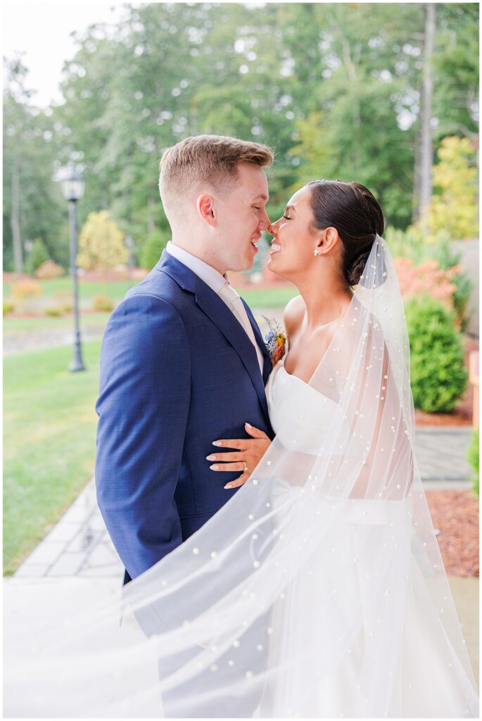 Bride and groom standing close together, smiling with the bride’s veil flowing around them, outside Pembroke Pines Country Club in Pembroke, NH.