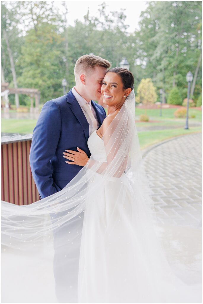 Bride and groom smiling at each other as the groom whispers into the bride’s ear, with the bride’s veil softly draped around them at Pembroke Pines Country Club in Pembroke, NH.