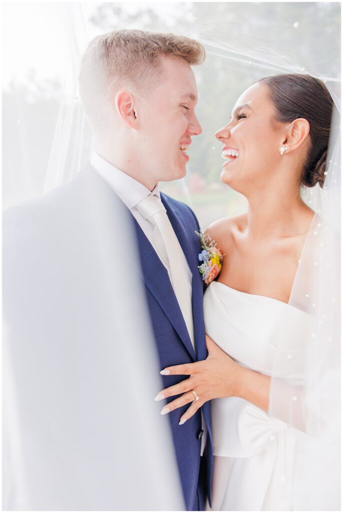 Bride and groom laughing together with the bride’s veil wrapped around them, standing outside at Pembroke Pines Country Club in Pembroke, NH.