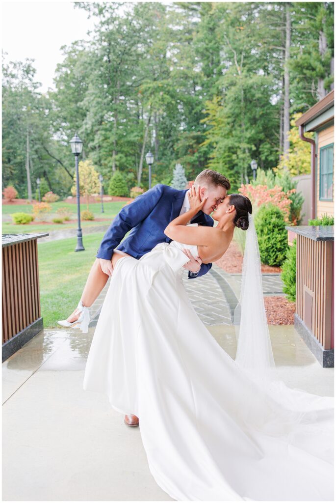 The groom dips the bride for a kiss outside under the covered patio at Pembroke Pines Country Club in Pembroke, NH, with a green, tree-lined background.