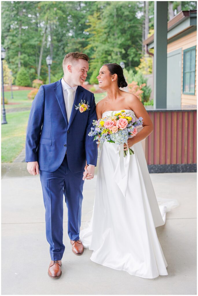 Bride and groom walking hand in hand, smiling at each other under the covered patio at Pembroke Pines Country Club in Pembroke, NH, with the bride holding a colorful bouquet.