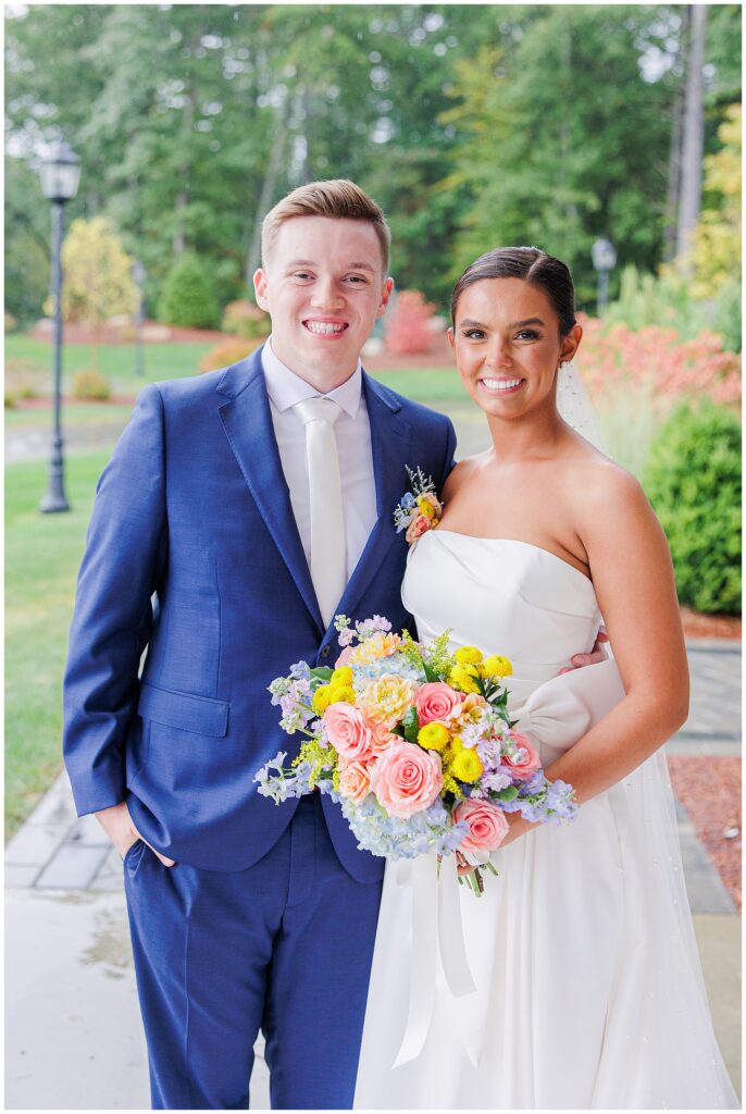 Bride and groom posing outside at Pembroke Pines Country Club in Pembroke, NH, both smiling at the camera with a green garden landscape behind them.