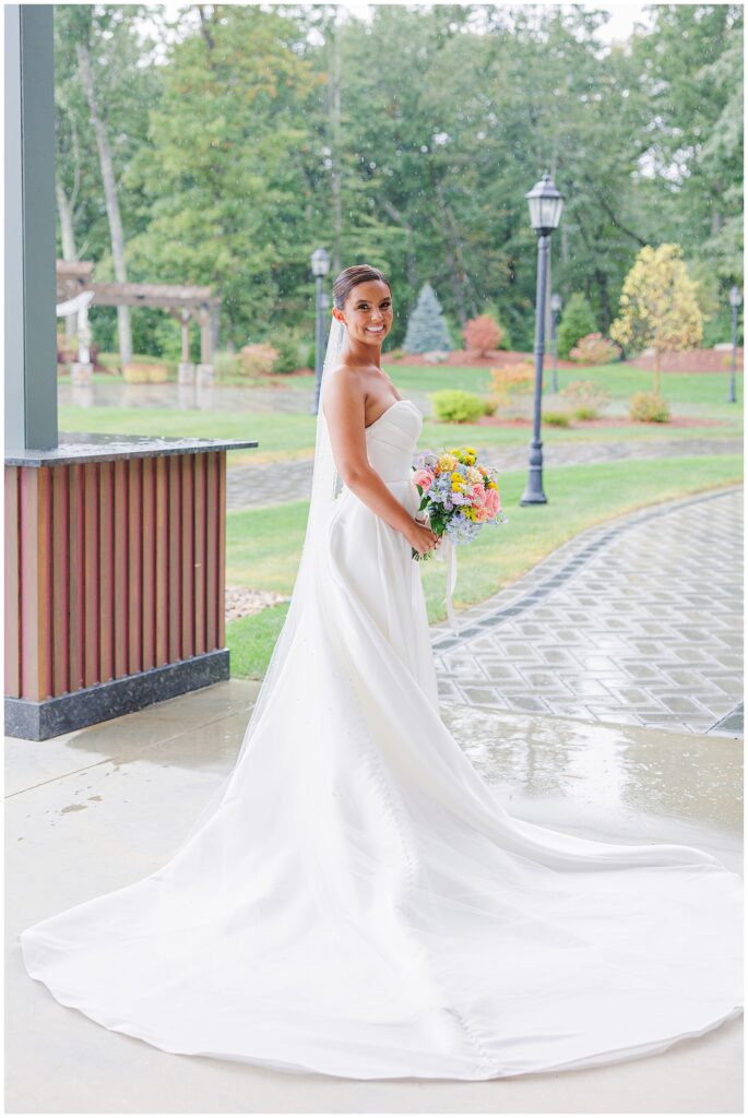 Full-length shot of the bride in her white dress, holding her bouquet and smiling outside at Pembroke Pines Country Club in Pembroke, NH, with a rain-soaked garden and trees in the background.