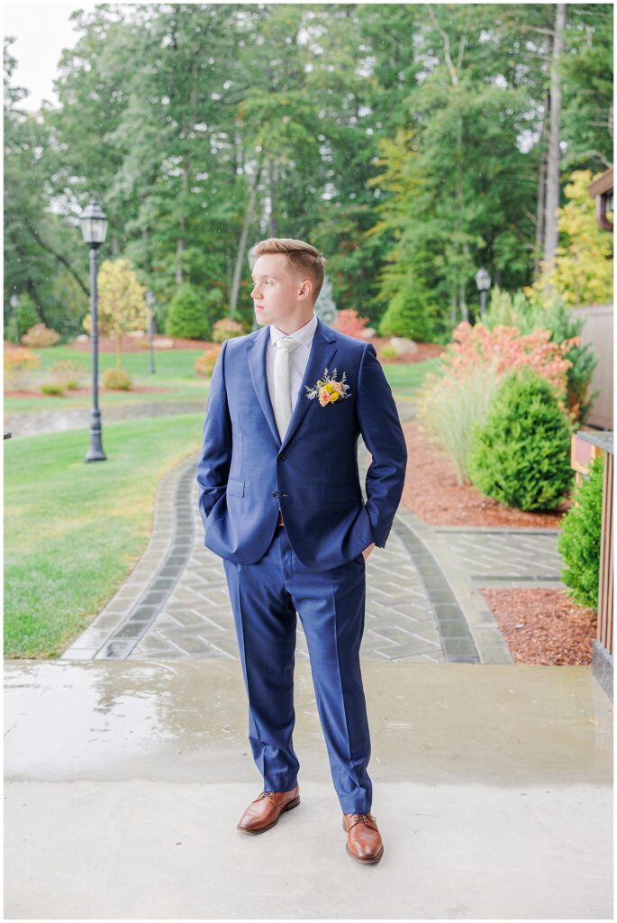 The groom standing outside at Pembroke Pines Country Club in Pembroke, NH, wearing a blue suit and gazing off to the side, with a scenic green landscape behind him.