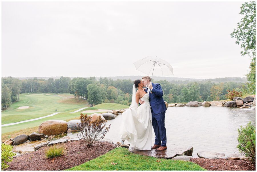 Bride and groom kissing under a clear umbrella near a pond with a view of the golf course at Pembroke Pines Country Club in Pembroke, NH, on a rainy day.