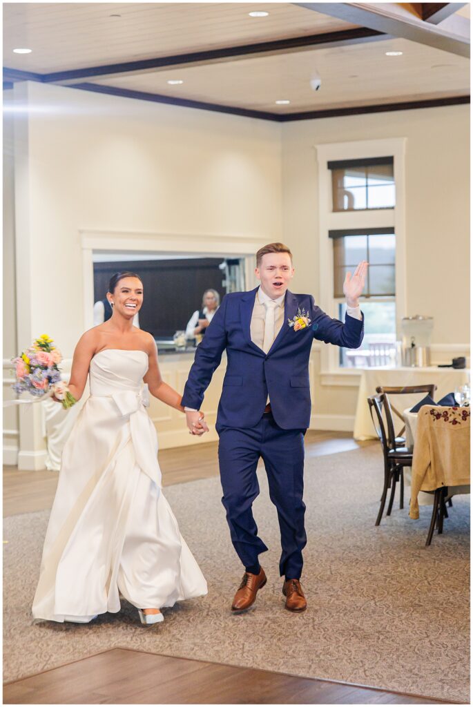 Bride and groom making their reception entrance at Pembroke Pines Country Club in Pembroke, NH, holding hands and smiling as the groom waves excitedly.