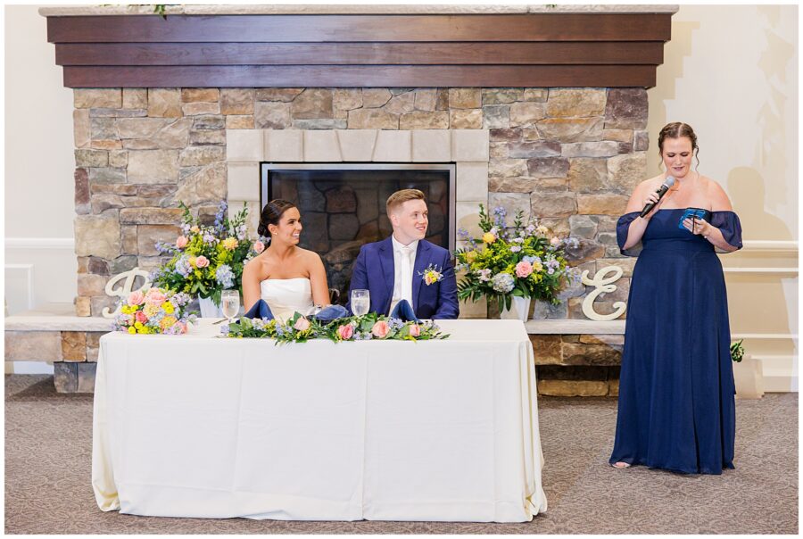 The maid of honor giving a speech while the bride and groom sit at the sweetheart table in front of a stone fireplace at Pembroke Pines Country Club in Pembroke, NH.