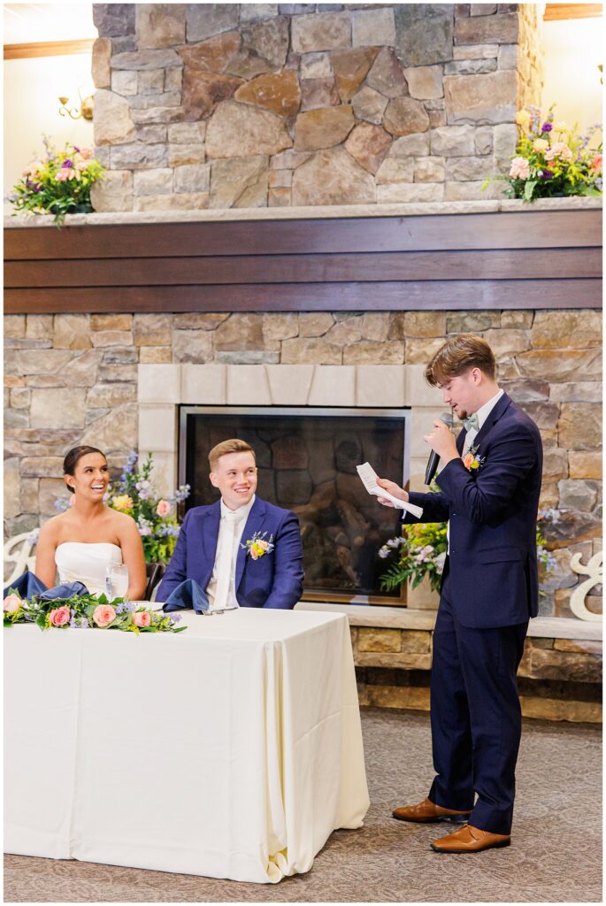 The best man delivers a speech while the bride and groom smile, seated in front of a stone fireplace at Pembroke Pines Country Club in Pembroke, NH.