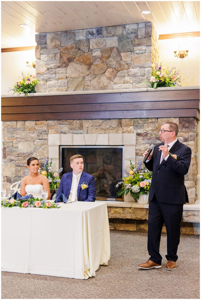 A guest gives a speech during the wedding reception at Pembroke Pines Country Club in Pembroke, NH, with the bride and groom seated in front of a decorated stone fireplace.