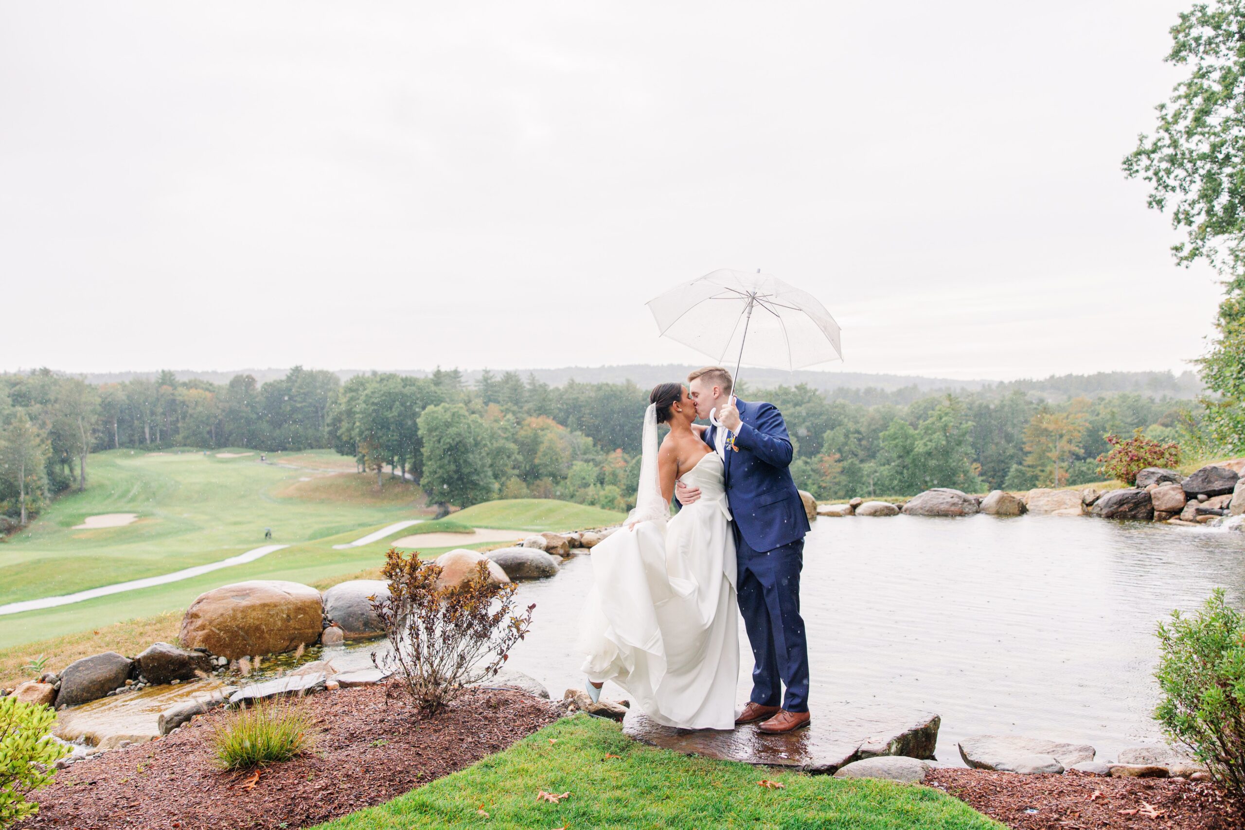 Bride and groom kissing under a clear umbrella near a pond with a view of the golf course at Pembroke Pines Country Club in Pembroke, NH, on a rainy day.