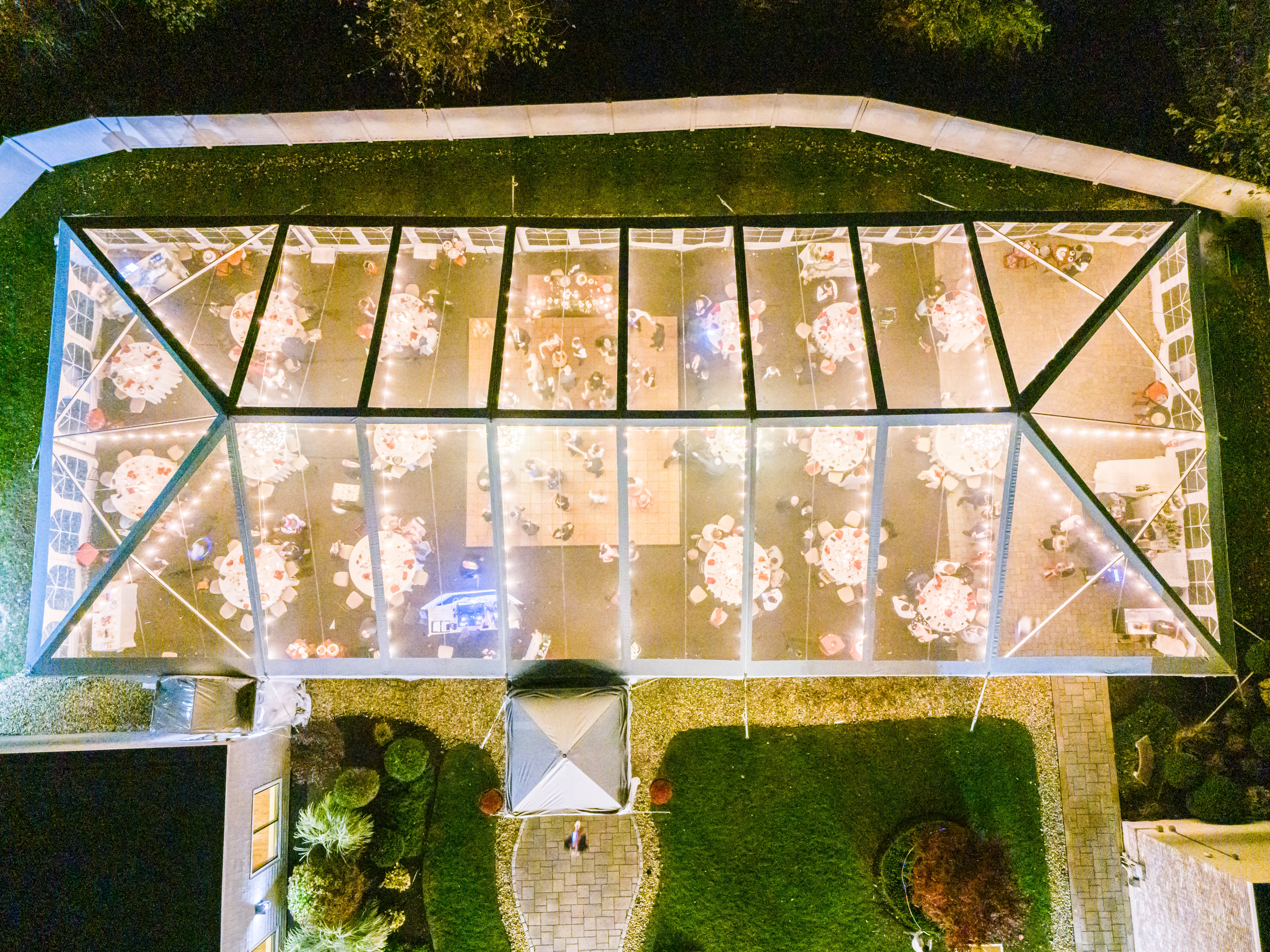 An overhead night drone shot of a large clear tent set up at the Colonial Hotel for a wedding reception. Inside the tent, tables with white tablecloths and red accents are arranged around a dance floor, with warm string lights creating a cozy ambiance.