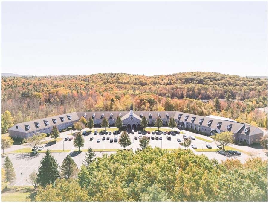 Aerial view of the Colonial Hotel in Gardner, MA – The Colonial Hotel, a long U-shaped building with dormer windows along the roof, is surrounded by colorful fall foliage. The hotel parking lot in front is filled with cars, and trees in vibrant shades of red, orange, and green extend into the distance.