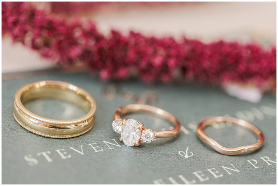 Wedding ring set display – Three rings, including a plain gold wedding band, a diamond engagement ring with side stones, and a gold wedding band with a wavy design, are displayed on a dark green surface with the faint text “Steven & Eileen.” Pink autumn foliage is in the background.