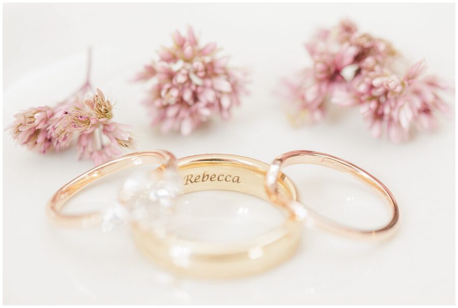 Engraved wedding band with flowers – A gold wedding band engraved with the name “Rebecca” is shown alongside two other wedding rings on a white surface. Small pink flowers are arranged behind the rings.