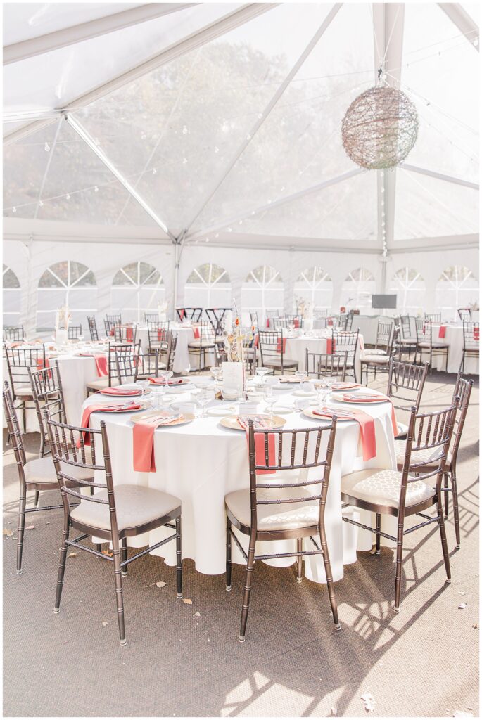 Outdoor wedding reception tent – Round tables set for a wedding reception inside a clear tent, each table featuring white tablecloths, gold-rimmed plates, red napkins, and simple glassware. An overhead wire ball decor light hangs from the tent ceiling, with soft natural light filtering through.