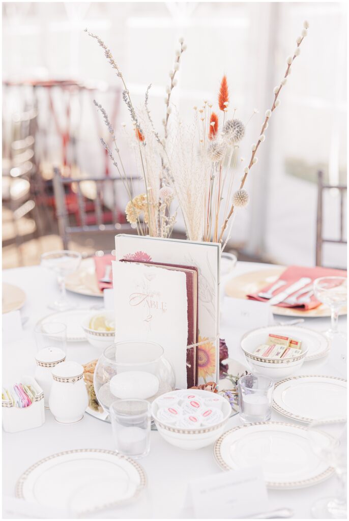 Table centerpiece with fall-inspired decor – A close-up of a table centerpiece featuring dried flowers in neutral tones with pops of red and orange, placed in a small glass vase on a wedding reception table.