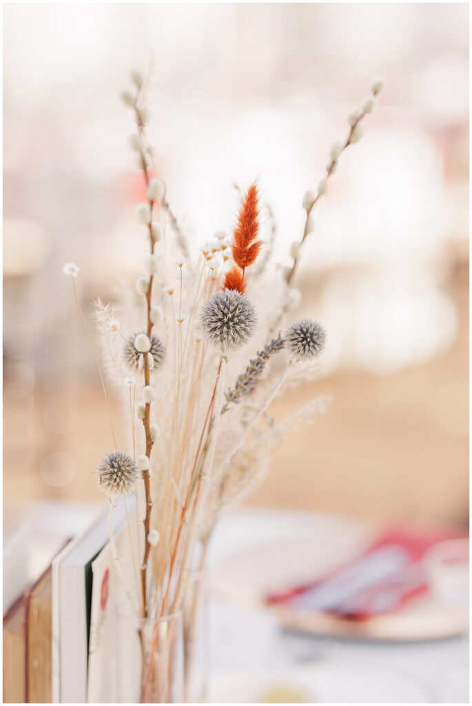 Close-up of dried floral centerpiece – A tall arrangement of dried flowers in natural tones, with accents of soft red and orange, sits in a clear vase as part of a wedding table centerpiece.