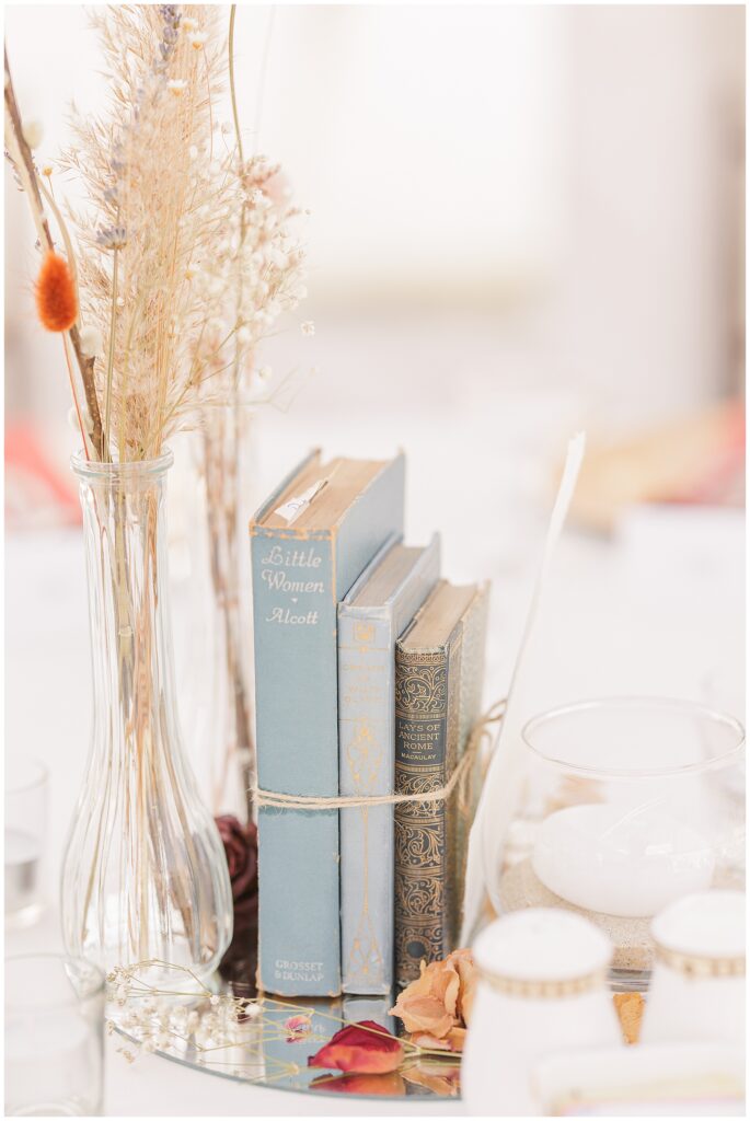 Vintage book centerpiece – A stack of vintage books, including “Little Women” by Alcott, tied together with twine and set alongside dried flowers and a glass candle holder, is displayed on a reflective surface as a wedding table centerpiece.